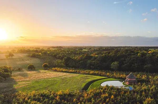 Aerial view of a thatched roundhouse and wild swimming pond at sunset over the estate